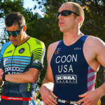 Two athletes in triathlon gear stand outdoors. One checks his watch while the other looks ahead. Sunny day with trees in the background.