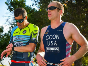 Two athletes in triathlon gear stand outdoors. One checks his watch while the other looks ahead. Sunny day with trees in the background.