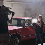 Film crew shoots a scene with a red pickup truck. A camera operator films while a person holds a clapperboard in front of the vehicle. Two people sit inside the truck.