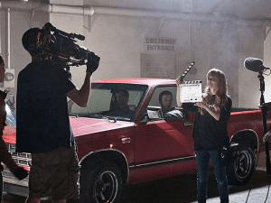 Film crew shoots a scene with a red pickup truck. A camera operator films while a person holds a clapperboard in front of the vehicle. Two people sit inside the truck.