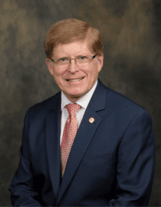 A person with glasses wearing a blue suit, white shirt, and red patterned tie is smiling against a dark, textured background.