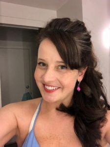 A woman with long brown hair smiles at the camera, wearing a blue top and pink earrings.