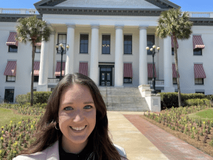 A person stands smiling in front of a large white building with columns and red-striped awnings.