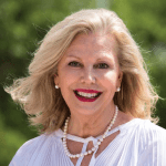 An older woman with shoulder-length blonde hair, wearing a white blouse and pearl necklace, smiles in an outdoor setting.