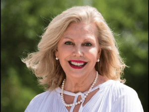 An older woman with shoulder-length blonde hair, wearing a white blouse and pearl necklace, smiles in an outdoor setting.