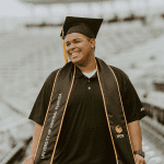 A graduate wearing a cap and gown stands in a stadium. The gown has "University of Central Florida" written on it. The person has a slight smile.