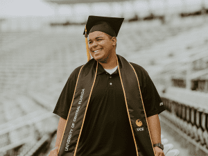 A graduate wearing a cap and gown stands in a stadium. The gown has "University of Central Florida" written on it. The person has a slight smile.