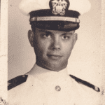 Portrait of a man in a white military uniform with a peaked cap featuring an emblem, against a plain backdrop.