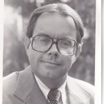 A man in a suit and tie with large glasses is smiling slightly, posed outdoors with a blurred natural background.