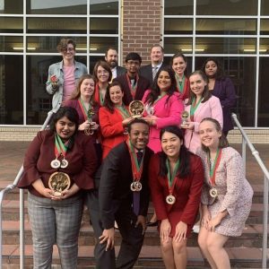 A group of people in formal attire smile on a staircase, each holding medals and trophies. Brick building and windows in the background.