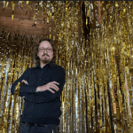 Man with long hair and glasses stands with arms crossed in front of gold foil streamers hanging from the ceiling.