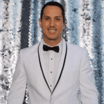 A man in a white tuxedo with a black bow tie stands in front of a silver sequin backdrop.