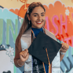 A graduate smiles, holding a cap, in front of a colorful background with motivational text. She's wearing a blue dress and a graduation stole with multiple cords.