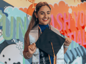 A graduate smiles, holding a cap, in front of a colorful background with motivational text. She's wearing a blue dress and a graduation stole with multiple cords.