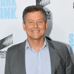 A man in a gray suit and blue shirt smiles at the "Shark Tank" event backdrop.