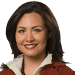 Portrait of a woman with dark hair, wearing a red blazer, white shirt, and a necklace, smiling at the camera against a plain white background.