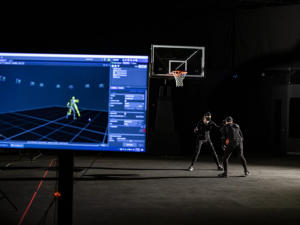 Two people in motion capture suits practice basketball moves on a court. A monitor displays their movements as digital models in real-time.