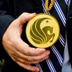 A person wearing a striped tie holds a large gold medallion with an embossed design featuring an abstract emblem.