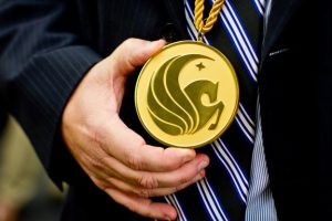 A person wearing a striped tie holds a large gold medallion with an embossed design featuring an abstract emblem.