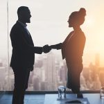 Two people in business attire shake hands in front of a window with a cityscape view and a bright sunset.