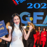 A woman in a white dress holds a trophy and certificate on stage at the 2021 Golden Film Award. A group of people stands in the background.