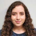 A woman with long, curly brown hair and light skin is facing the camera, wearing a dark patterned top and a thin necklace, with a neutral background.