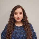 A person with long curly hair wearing a blue-patterned blouse stands against a neutral background.