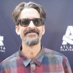 Man with sunglasses and a beard smiling at the camera, wearing a plaid shirt. Background shows a sign for Atlas Film Festival.