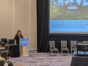 A person stands at a podium giving a presentation about the Virtual UCF Arboretum. A large screen displays slides related to the project. Audience members are seated.