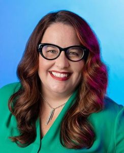 A person with long brown hair, wearing glasses, a green top, and a necklace, smiling against a blue background.