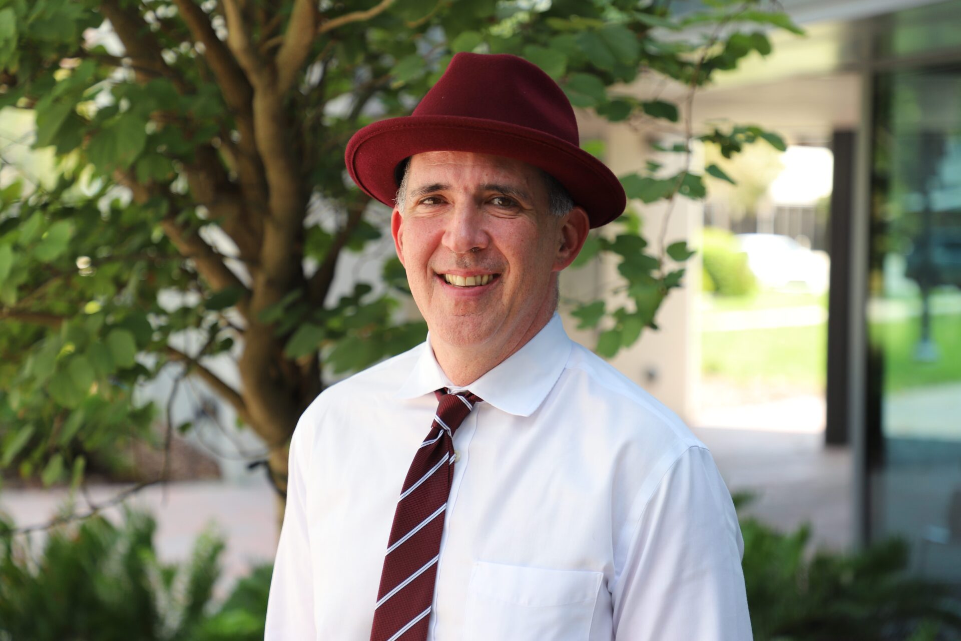 Man wearing a red hat, white shirt, and striped tie stands outside near a tree, smiling at the camera.