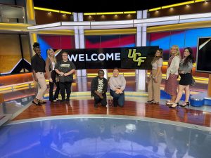 A group of people stands in front of a "Welcome UCF" sign in a brightly lit studio setting.