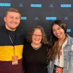 Three people smiling in front of a backdrop with the Enzian logo. Two wear event lanyards.