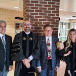 Four adults stand together indoors, two in academic regalia, next to a brick wall. Two of the adults hold toddlers.