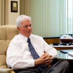 An elderly man in a white shirt and patterned tie sits in an office chair, hands clasped. A football and book are on the desk beside him. Vertical blinds cover a window in the background.