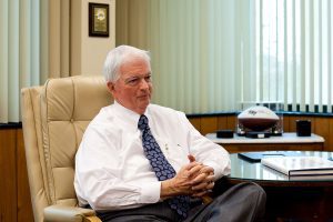 An elderly man in a white shirt and patterned tie sits in an office chair, hands clasped. A football and book are on the desk beside him. Vertical blinds cover a window in the background.