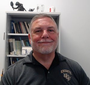 Man with a gray beard smiles at the camera, wearing a "Knights UCF" polo shirt. Bookshelves with various items are visible in the background.