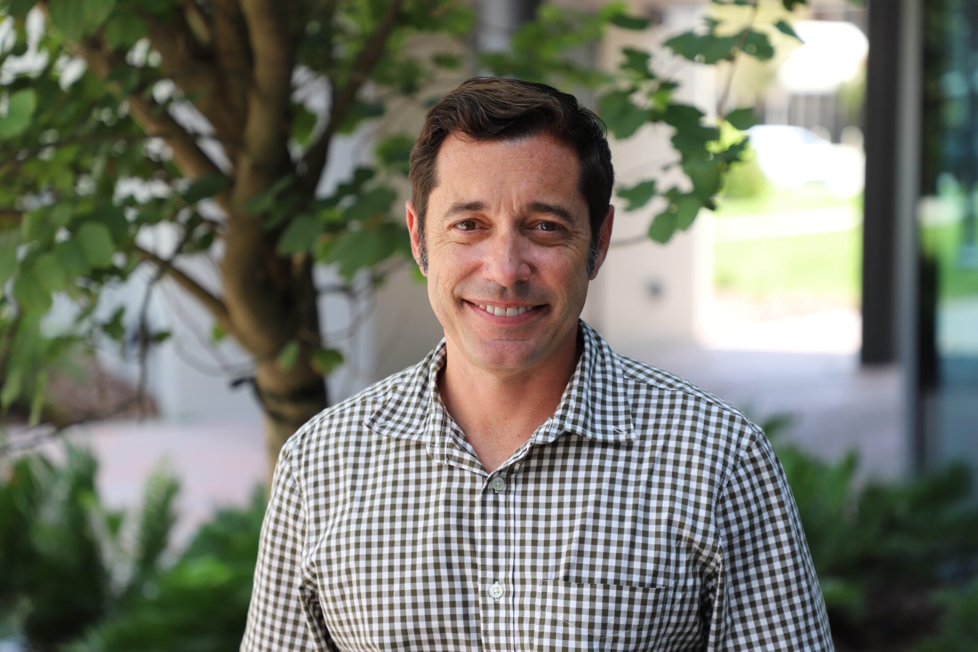 Man in a checkered shirt stands outdoors in front of a tree and greenery, smiling at the camera.