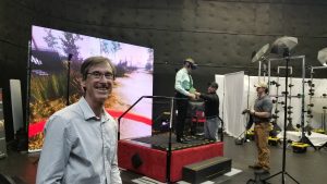 A man smiles at the camera in a virtual reality studio. In the background, two men interact with VR equipment on a platform. A large screen displays a scenic virtual environment.