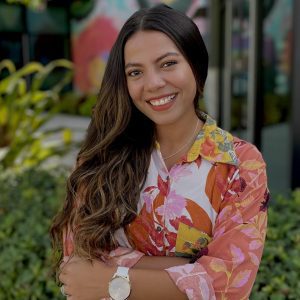 Smiling person with long hair wearing a floral shirt stands outdoors with greenery and a colorful mural in the background.