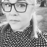 Black and white close-up of a person wearing glasses and a patterned top, standing outdoors with grass and trees in the background.