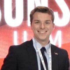 A person in formal attire smiles while standing in front of a blurred background with large white and red text.