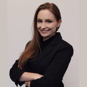 A woman in a black blazer stands with arms crossed, smiling against a plain background.