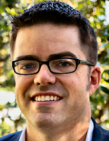 A person with short dark hair and glasses smiles outdoors with greenery in the background.