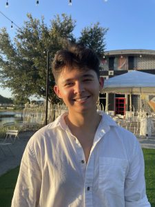 A person in a white shirt smiles outdoors, standing on grass with a background of trees and a building under a clear blue sky.