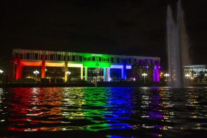 A building is illuminated with rainbow colors at night, reflected in a body of water. A fountain sprays water in the foreground.