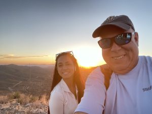 Two people take a selfie on a hilltop at sunset, with winding roads and hills in the background. One wears a cap and sunglasses, and both are smiling.