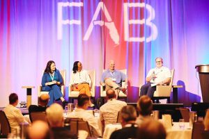 Four panelists sit on stage discussing at a conference with an audience. The backdrop displays large letters "FAB.