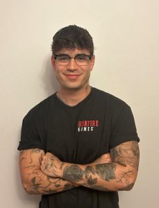 A person with tattoos on their arms and wearing glasses is smiling, standing with arms crossed. They are wearing a black T-shirt with "Gunfire Games" written on it.