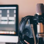 Close-up of a professional microphone and headphones in a recording studio. A computer screen with audio editing software is visible in the blurred background.